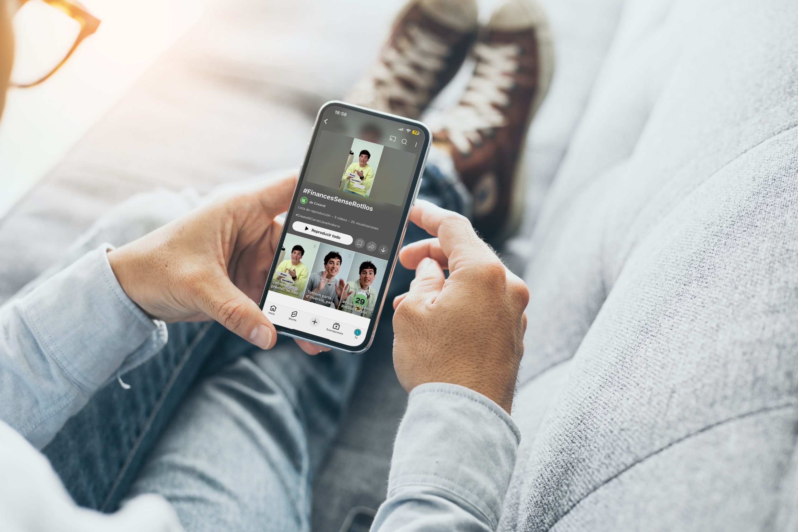 JovesCreand 6 Persona asseguda en un sofà, sostenint un mòbil que mostra un perfil de xarxa social amb el títol #FinancesSenseRotllos i diverses miniatures de vídeo.
