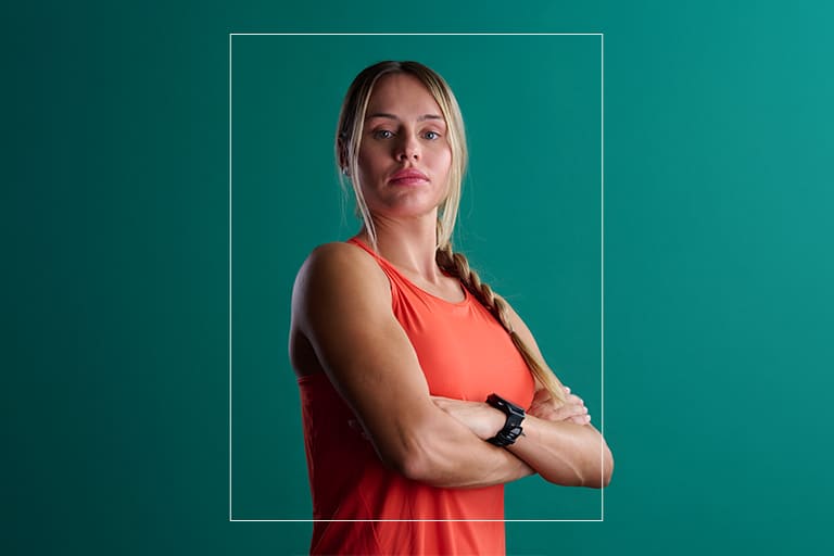 JovesCreand 5 Retrat d'una dona en roba esportiva taronja, amb els braços plegats i un rellotge intel·ligent, sobre un fons verd fosc.